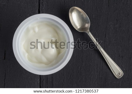 Yogurt in plastic cup on rustic black table with small silver spoon - natural Greek yoghurt top view photo Royalty-Free Stock Photo #1217380537