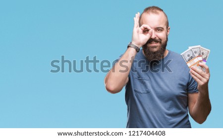 Young hipster man holding bunch of money over isolated background with happy face smiling doing ok sign with hand on eye looking through fingers