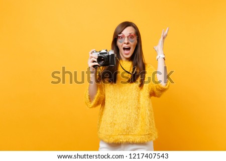 Excited young woman in heart eyeglasses spreading hand taking pictures on retro vintage photo camera isolated on bright yellow background. People sincere emotions, lifestyle concept. Advertising area
