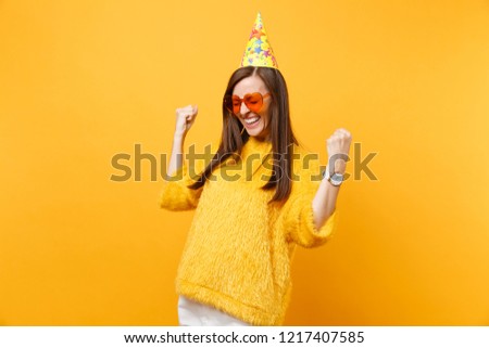 Joyful happy young woman in orange heart glasses, birthday party hat clenching fists like winner enjoying holiday, celebrating isolated on bright yellow background. People sincere emotions, lifestyle