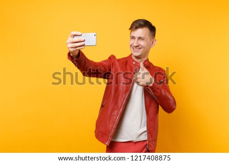 Portrait handsome young man in red leather jacket, t-shirt doing selfie on mobile phone isolated on bright trending yellow background. People sincere emotions lifestyle concept. Advertising area