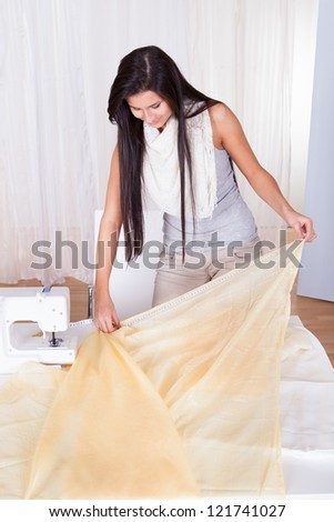 Attractive stylish seamstress or interior designer measuring a length of neutral colored fabric alongside her sewing machine with copyspace