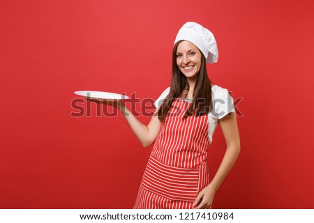 Housewife female chef cook or baker in striped apron white t-shirt, toque chefs hat isolated on red wall background. Woman hold empty blank round plate with place for food. Mock up copy space concept