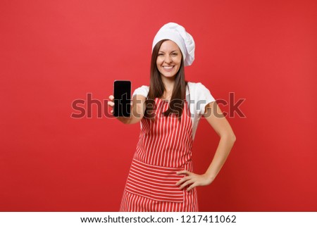 Housewife female chef cook or baker in striped apron t-shirt toque chefs hat isolated on red wall background. Woman hold cellphone with blank screen for promotional content. Mock up copy space concept