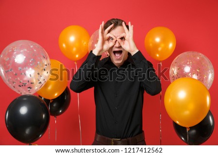 Funny young man in black classic shirt showing OK sign near eyes on bright red background air balloons. St. Valentine's, International Women's Day Happy New Year birthday mockup holiday party concept