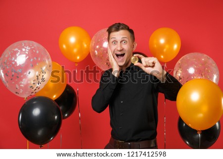Excited young man in classic shirt keeping hand on cheek holding bitcoin metal coin of golden color future currency on red background air balloon. Happy New Year birthday mockup holiday party concept