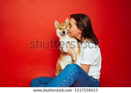 Cute brunette woman in white t shirt and jeans holding and embracing Shiba Inu dog on plane red background. Love to the animals, pets concept Royalty-Free Stock Photo #1217558653