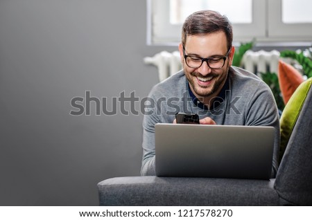 Handsome freelancer working from home, lying on his stomach on the couch with laptop and phone. Smiling. Wearing glasses. Copy space.  Royalty-Free Stock Photo #1217578270