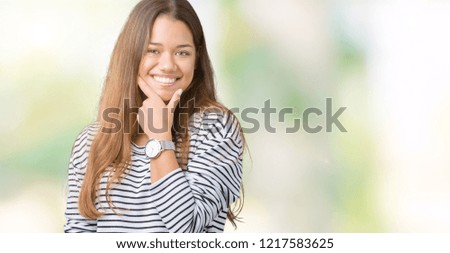Young beautiful brunette woman wearing stripes sweater over isolated background looking confident at the camera with smile with crossed arms and hand raised on chin. Thinking positive.