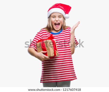 Young beautiful girl wearing christmas hat and holding gift over isolated background very happy and excited, winner expression celebrating victory screaming with big smile and raised hands