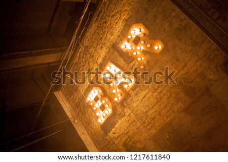 Luminous letters BAR. Volumetric letters BAR from rusty metal on a brick wall with a garland of incandescent lamps. BAR signboard. Inscription BAR from large metal letters decorated with glowing light