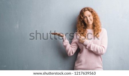Young redhead woman over grey grunge wall wearing pink sweater amazed and smiling to the camera while presenting with hand and pointing with finger.
