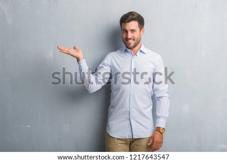 Handsome young business man over grey grunge wall wearing elegant shirt smiling cheerful presenting and pointing with palm of hand looking at the camera.