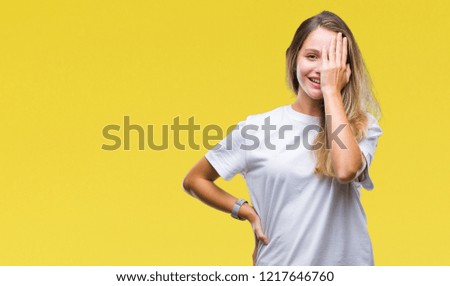 Young beautiful blonde woman wearing casual white t-shirt over isolated background covering one eye with hand with confident smile on face and surprise emotion.