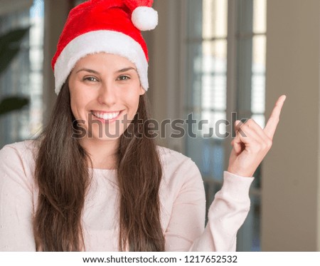 Beautiful young woman wearing Santa Claus hat at home with a big smile on face, pointing with hand and finger to the side looking at the camera.