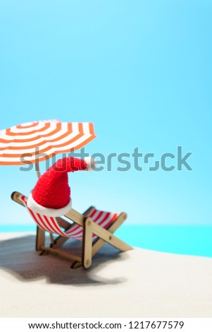 Cartoon arrangement of Santa hat on deck chair under sunshade on beach against blue background