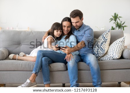 Multi-ethnic family sitting on sofa in living room at home. Mother father and little daughter using smartphone watching recreational program funny videos online having videocall talking with friends