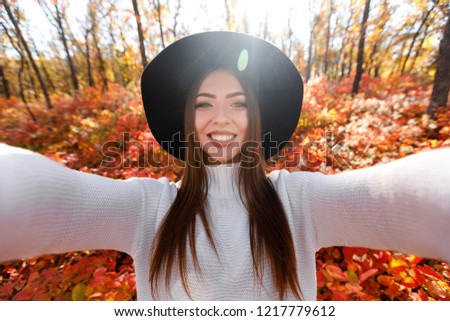 Portrait of beautiful young happy woman in black hat making selfie in the autumn park full of yellow leaves