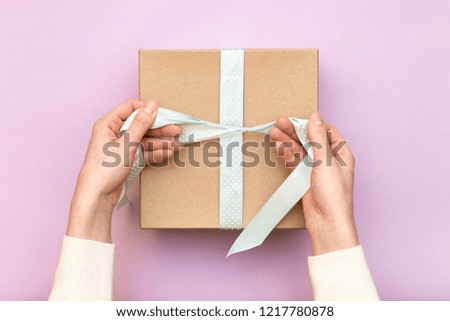 Woman hands holding craft gift box with blue bow on pastel pink background. Girl binds classic bow for present box. Top view, flat lay style, copy space