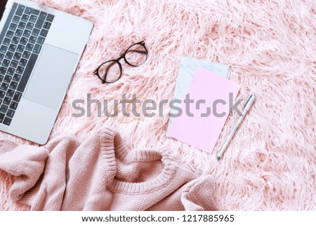 Flatlay of laptop, women knitted sweater, reading glasses, paper, pen on a pink fur background