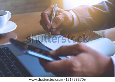 Businessman hand holding smart phone  , writing and signing checkbook, work on laptop computer on wooden table at modern office. Paycheck concept. 