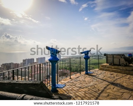 A close up photo of two blue public monocular and cannons with spectacular aerial views of the Mediterranean Sea and the seaside touristic town of Cullera, which is located in Valencia, Spain, Europe.