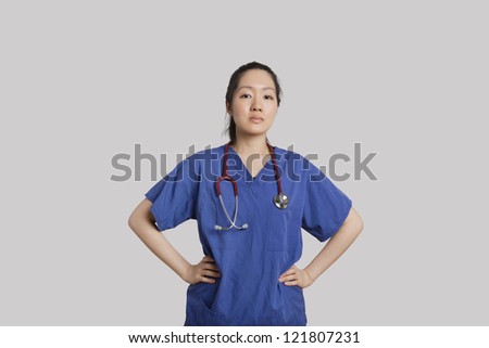 Portrait of a confident young Asian female doctor standing with hands on hips over gray background