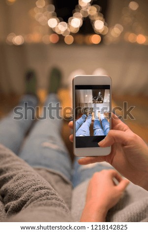 leisure, hygge and christmas concept - close up of couple taking foot selfie by smartphone and garland lights at home
