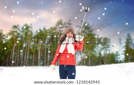 people, technology and leisure concept - happy woman in winter fur hat taking picture by smartphone selfie stick over winter forest background and show