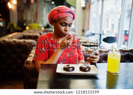 Stylish african woman in red shirt and hat posed indoor cafe, eat chocolate dessert cakes and looking at mobile phone.