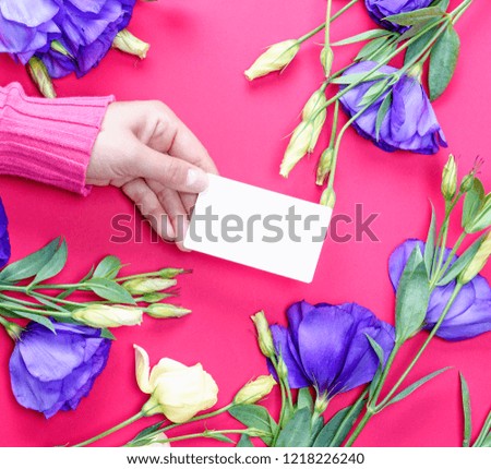 female hand in pink sweater holding a blank white paper business card on a pink background with flowers Eustoma Lisianthus