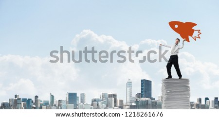 Ambitious and young businessman in suit throwing big drawn rocket from hand while standing on paper column with cityscape view on background