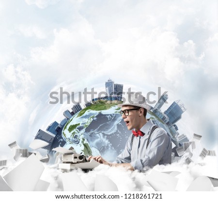 Emotional young man writer in hat and eyeglasses feeling surprised while using typing machine at the table with flying papers and Earth globe among cloudy skyscape on background. Elements of this