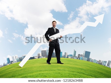 Conceptual image of young successful business man in black suit pointing aside by means of big white banner in form of arrow while standing on green meadow with city on background.