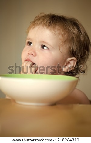 Portrait of cute curious small boy with blonde curly hair and round cheecks eating from green plate with hand closeup, vertical picture