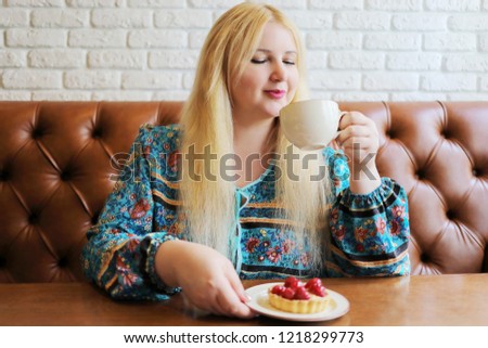 Pretty fat blonde drinks tea in cafe, cake with berries is on table
