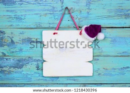 Blank white wood sign hanging by ribbon on antique rustic teal blue wood door with purple Christmas Santa Claus hat; holiday background with painted copy space