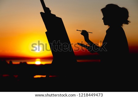 silhouette of a young woman painting a picture with paints on canvas on an easel, girl profile with paint brush and palette engaged in art on the nature in a field at sunset