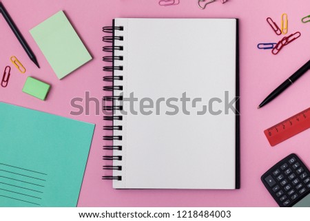 Empty spiral notebook with white page and multicolored stationery on a pink table. Workplace with notepad on colored background. Sketch-book for input the text, top view. Back to school concept.