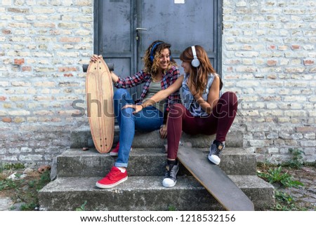 Two girl friends with headphones sitting at the stairs in front of metal door with skateboards and making fun