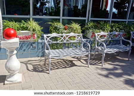 two gray metal benches on the sidewalk against the wall with windows and flowerpots with plants and flowers