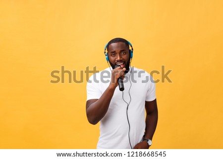 Portrait of cheerful positive chic handsome african man holding microphone and having headphones on head listening music singing song enjoying weekend vacation isolated on yellow background