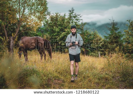 stylish bearded man in a hat and a gray jacket photographing nature among the ouu in the mountains, against the background of a blurred horse and evening slopes of the Carpathian mountains
