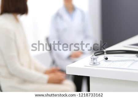 Stethoscope, clipboard with medical form lying on hospital reception desk with laptop computer and busy doctor and patient communicating at the background. Medical tools at doctor working table