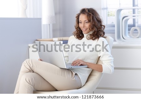 Pretty young woman using laptop computer at home, sitting in modern chair, smiling.