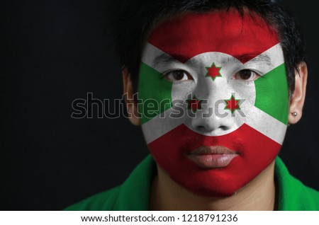 Portrait of a man with the flag of the Burundi painted on his face on black background, A white diagonal cross divided into four panels of red and green and three star.