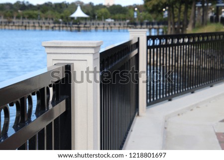 Perspective photography  of black metal fence with concrete posts and brick paver bridge over turquoise blue reflecting water lake.