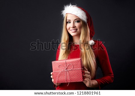 New Year, Christmas, x-mas, holidays, gifts and happiness concept - Happy content female shopper, woman in Santa helper hat with red gift box over black wall with blank space for promotion text