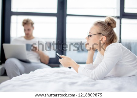 young woman is lying on the bed and choosing the chanel to watch while her boyfriend drinking tea and looking at the screen of the notebook. blurred background. close up side view photo