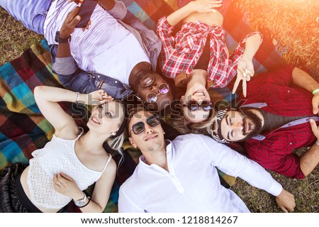 Multiethnic adult friends in casual summer wear lying in circle on the colourful rug in the park having fun enjoying togetherness and friendship.